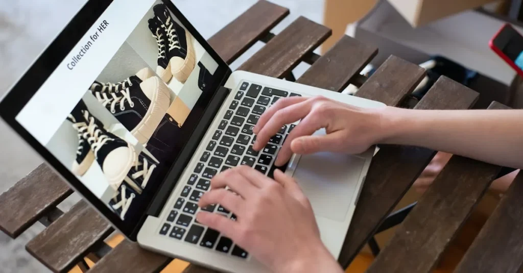 Person typing on a laptop displaying a webpage with images of black high-top sneakers labeled "Collection for HER," on a wooden table.