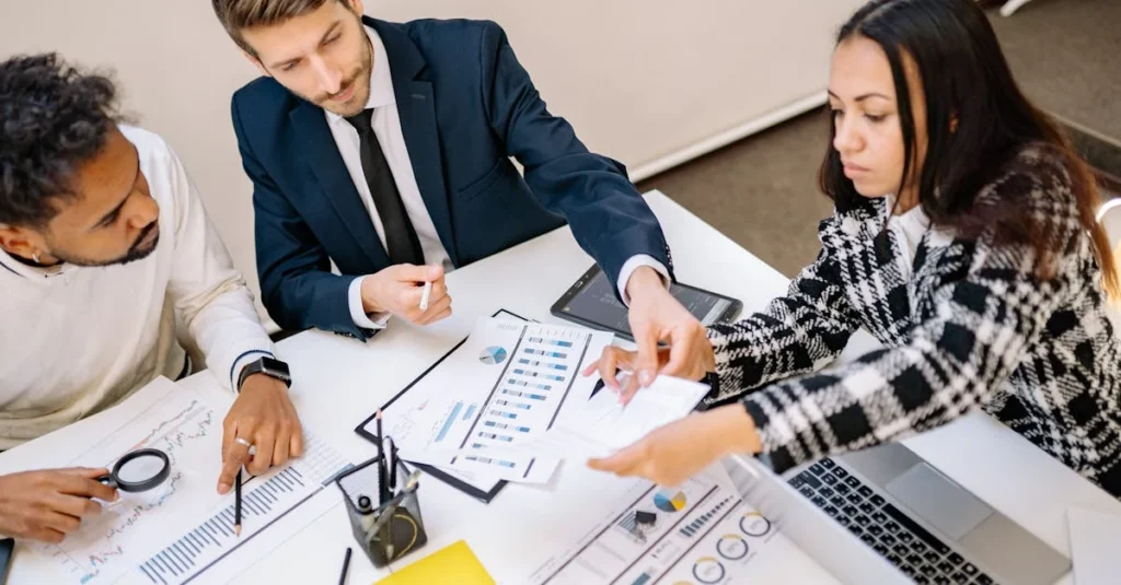 Three people collaborate at a table with charts and tablets, discussing data.