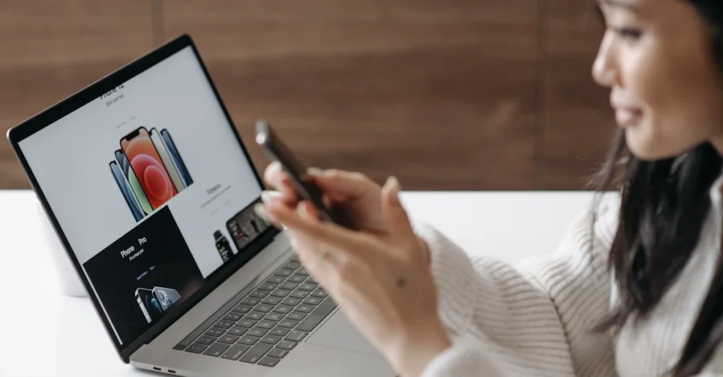 Woman using laptop displaying product catalog while holding smartphone.