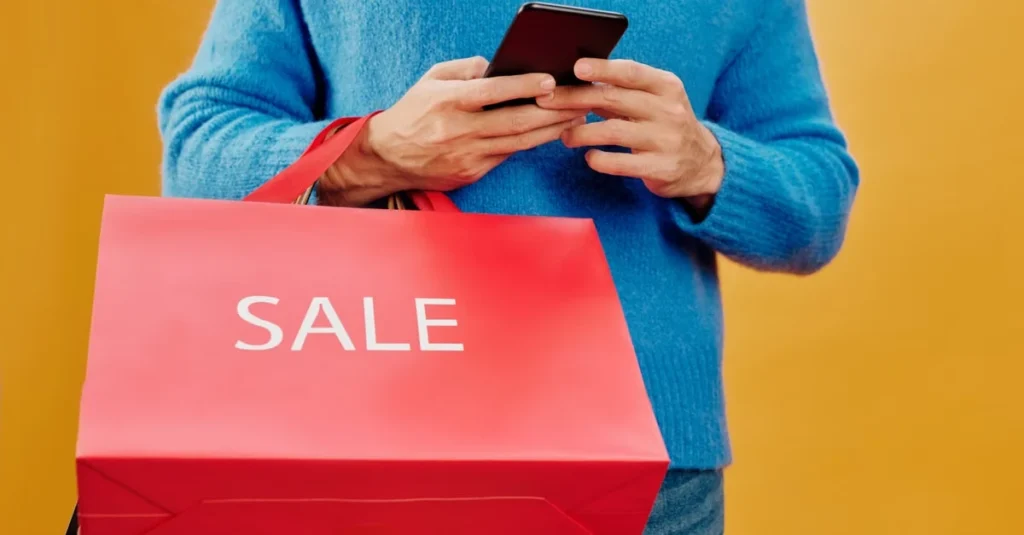 Person in blue sweater holding phone while carrying red SALE shopping bag against yellow background.