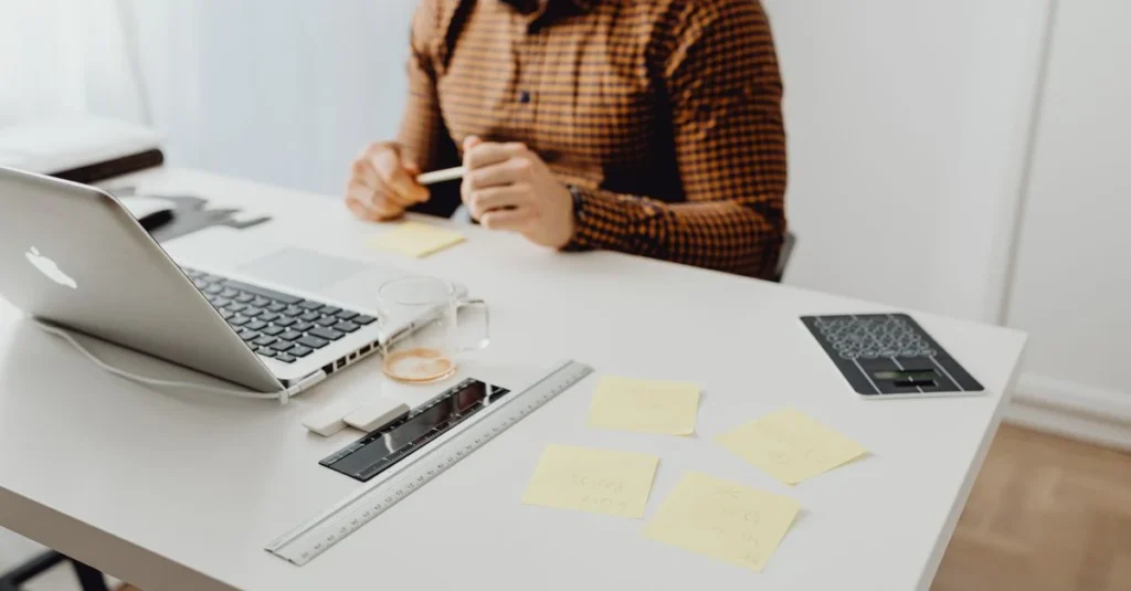 Woman in brown checkered shirt taking Nosto reviews notes at desk with laptop and sticky notes.