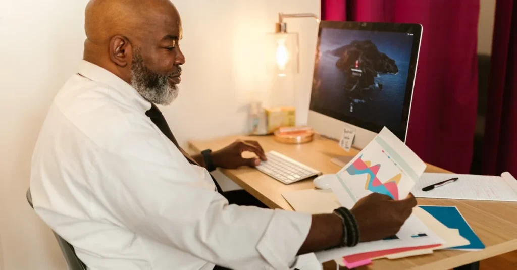 Man working at desk with computer monitor displaying interface, reviewing analytics and design materials.